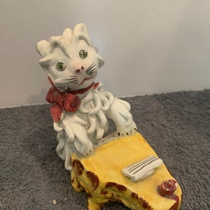 May include: A white ceramic cat figurine playing a yellow piano with a red bow. The cat has green eyes and black whiskers.