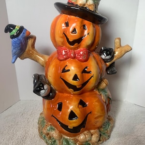 Könnte beinhalten: Keramik-Halloween-Dekoration mit drei gestapelten orangefarbenen Kürbissen mit lächelnden Gesichtern. Sie hat einen schwarzen Zylinder, eine rote Fliege und einen blauen Vogel und eine schwarze Katze auf Ästen. Der Sockel ist mit Blättern und Kürbissen verziert.
