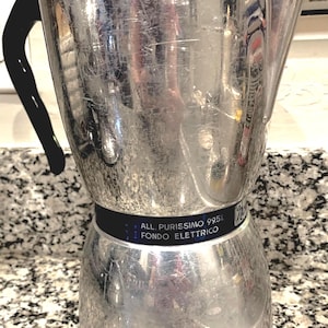 Può includere: Macchina per caffè espresso in argento con manico e coperchio neri. La macchina ha un'etichetta blu e bianca con la scritta "ALL. PURISSIMO 99.5% FONDO ELETTRICO".