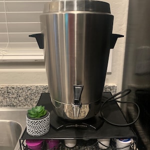 Può includere: Una caffettiera in acciaio inossidabile con base nera e cavo nero. La caffettiera è appoggiata su una griglia in metallo nero con una piccola pianta grassa in vaso davanti. La griglia ha diversi scomparti per le capsule di caffè.