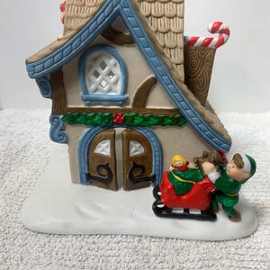Peut inclure: Une maison de village de Noël en céramique avec une base enneigée. La maison présente une bordure de toit bleue, des portes marron et une cheminée avec un sucre d'orge. Un elfe pousse un traîneau rouge avec des jouets.