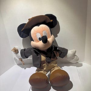 May include: A plush Mickey Mouse toy dressed in a brown leather jacket, khaki pants, and a brown hat. The toy has large black ears, a tan face, and white gloves. It is holding a small white object with a gold-colored rope.