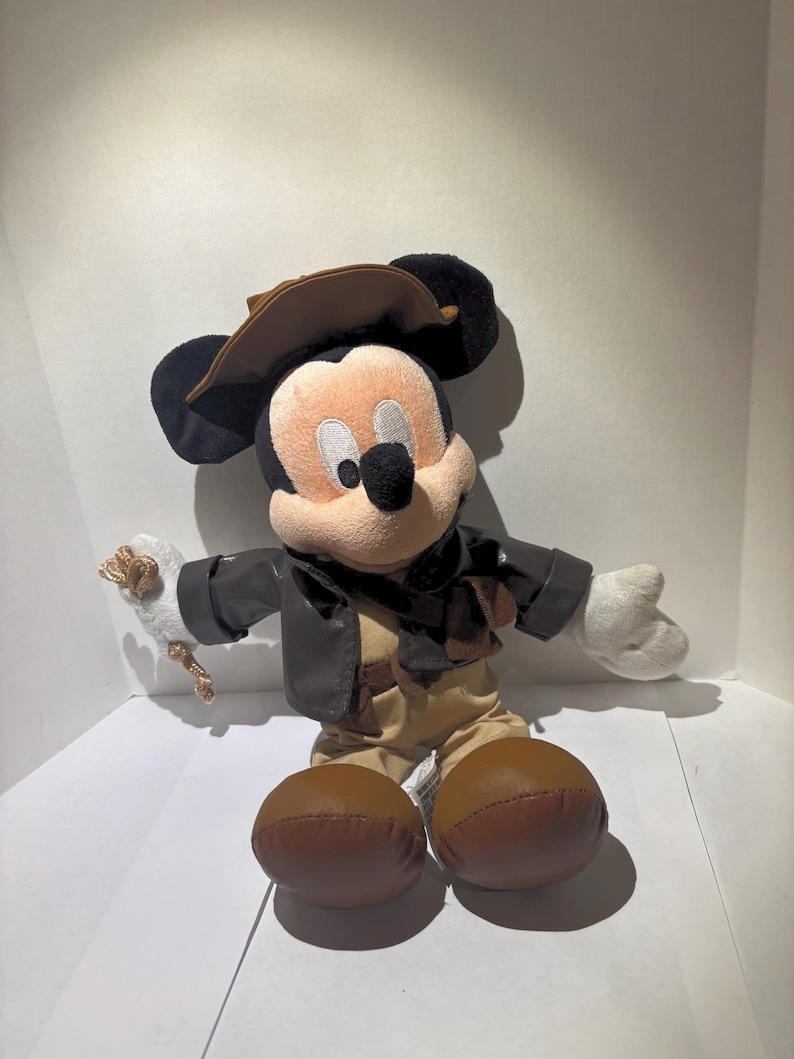 May include: A plush Mickey Mouse toy dressed in a brown leather jacket, khaki pants, and a brown hat. The toy has large black ears, a tan face, and white gloves. It is holding a small white object with a gold-colored rope.
