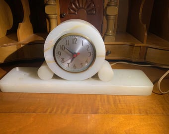 Könnte beinhalten: Eine weiße Alabasteruhr mit einem runden Zifferblatt und goldenen Zeigern. Die Uhr steht auf einem weißen Alabastersockel mit zwei abgerundeten Stützen.