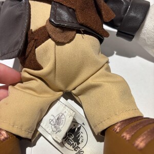 May include: A plush doll wearing a brown leather jacket, tan pants, and brown shoes. The doll has a brown scarf and a tag with the Disney logo. The doll's outfit is reminiscent of an adventurer.