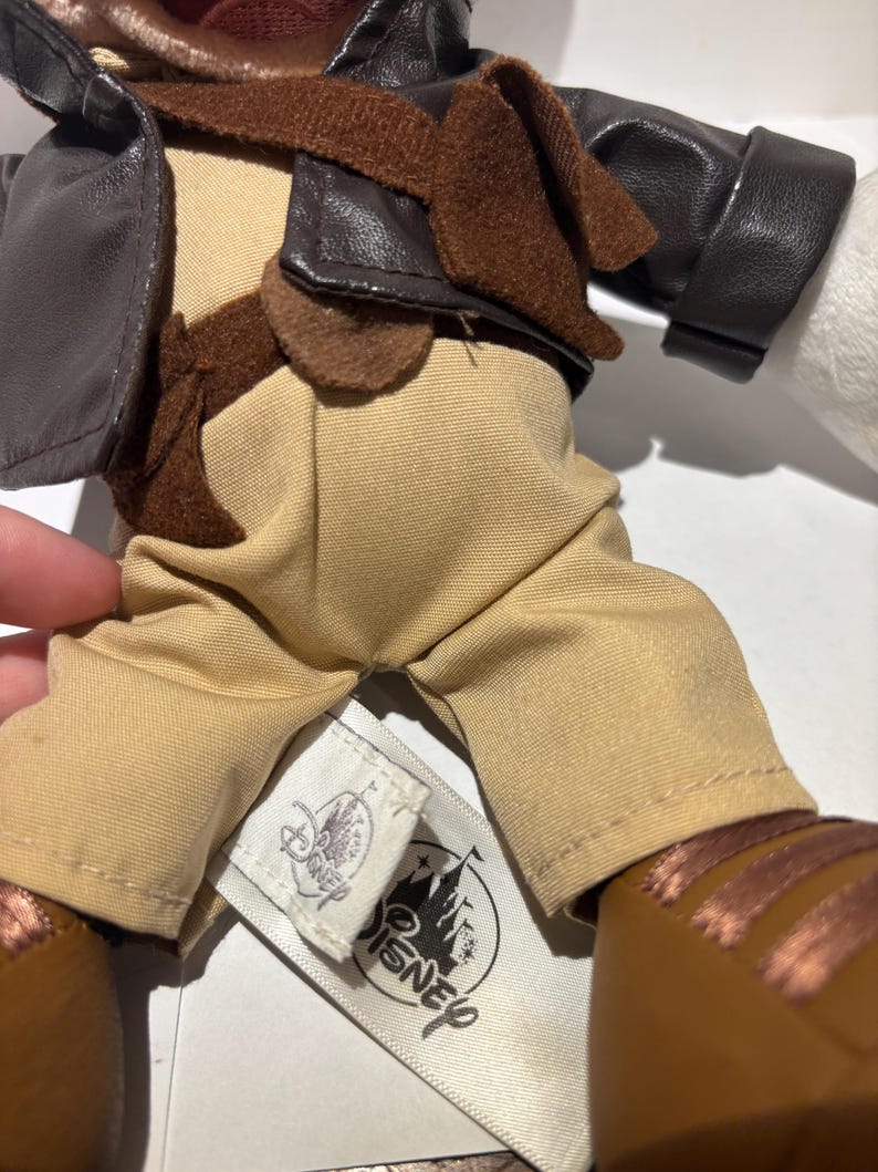May include: A plush doll wearing a brown leather jacket, tan pants, and brown shoes. The doll has a brown scarf and a tag with the Disney logo. The doll's outfit is reminiscent of an adventurer.