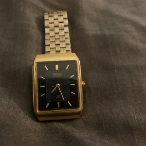 Puede incluir: Un reloj Seiko Quartz dorado con una esfera negra rectangular y manecillas e indicadores dorados. El reloj tiene una correa de eslabones dorados. La esfera del reloj muestra "SEIKO QUARTZ" y "JX".