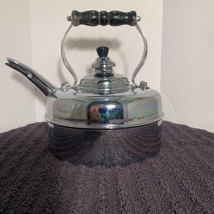 Può includere: Una lucida teiera cromata con manico e beccuccio in legno nero. La teiera ha un corpo arrotondato e un coperchio con un pomello nero. Il manico è arcuato e attaccato ai lati della teiera.