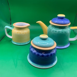 May include: A blue and green teapot, a yellow creamer, and a blue sugar bowl with a lid. The set is made of ceramic and has a hand-painted design.
