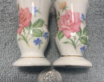 Puede incluir: Un par de saleros y pimenteros de cerámica blanca con un diseño floral. Los saleros tienen un borde dorado y presentan rosas rosas, flores azules y hojas verdes.