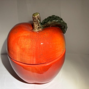Puede incluir: Un recipiente de cerámica con forma de manzana con tapa. El recipiente es principalmente rojo con reflejos anaranjados, y una hoja verde y un tallo marrón están unidos a la parte superior. El recipiente probablemente se utiliza para almacenamiento o decoración.