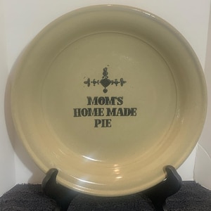 Puede incluir: Una fuente para pastel beige con el texto "MOM'S HOME MADE PIE" en letras negras. La fuente es redonda con un borde ligeramente elevado y está sobre un soporte negro.