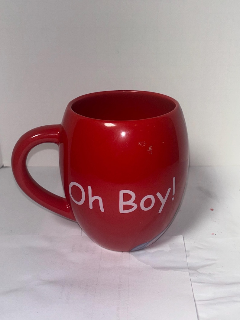May include: A glossy red ceramic mug with a rounded shape and a curved handle. The mug features the white text "Oh Boy!" on its side. The mug is designed for hot beverages and is suitable for home or office use.