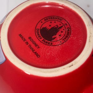 May include: Close-up of the bottom of a red ceramic mug. The mug's base features a white rim and a circular stamp with the text "AUTHENTIC DISNEY PARKS ORIGINAL" around a silhouette of a castle. The text "MADE IN THAILAND" is also visible.