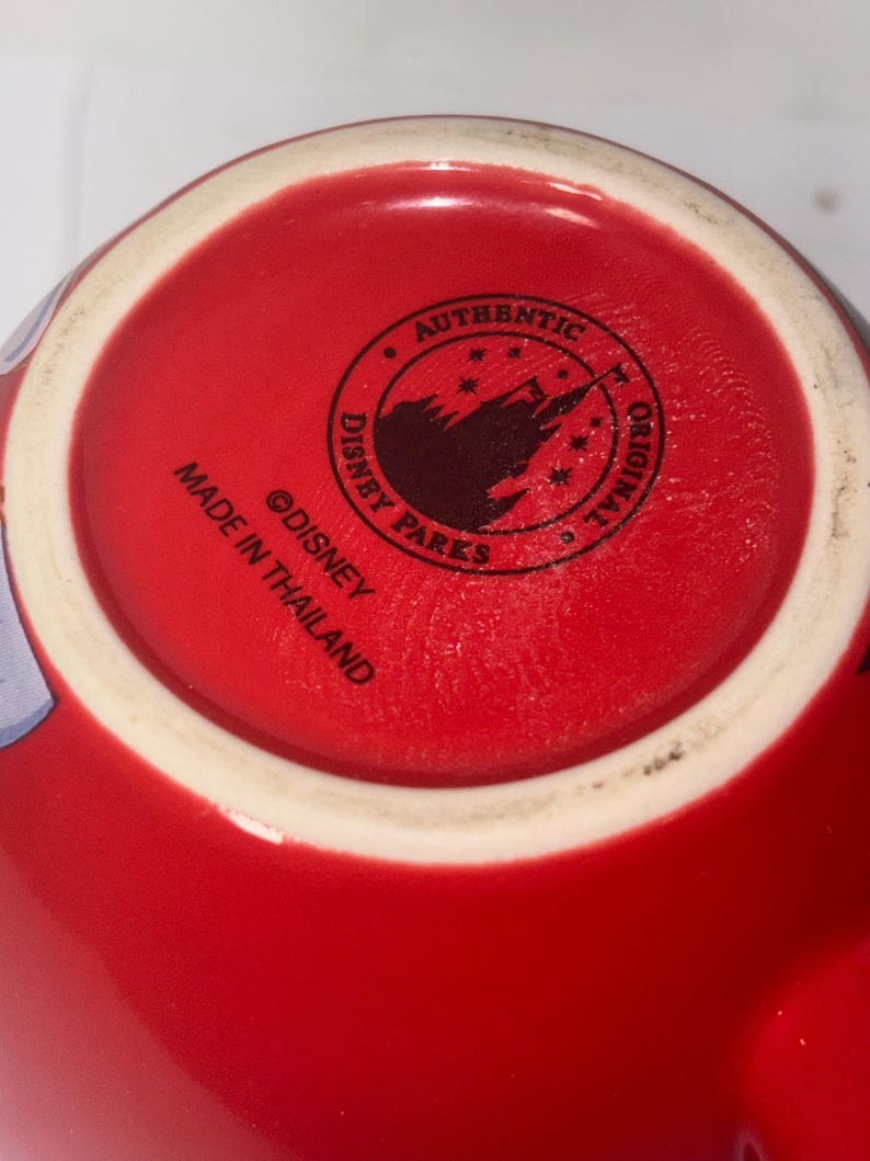 May include: Close-up of the bottom of a red ceramic mug. The mug's base features a white rim and a circular stamp with the text "AUTHENTIC DISNEY PARKS ORIGINAL" around a silhouette of a castle. The text "MADE IN THAILAND" is also visible.