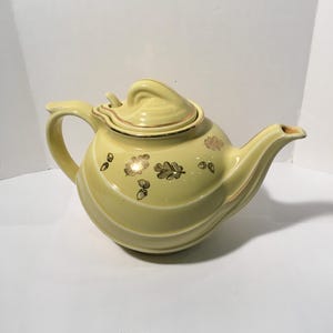 May include: A vintage yellow teapot with a matching lid. The teapot features a curved handle and spout, with gold leaf and acorn designs. The lid has a swirl design. The teapot is a light yellow color.