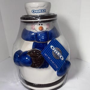 Op de afbeelding: Een keramische Oreo sneeuwman koekjespot. De pot is wit met een blauwe sjaal, handschoenen en hoed. De sneeuwman houdt een Oreo-koekje vast. De pot heeft het Oreo-logo op de hoed en een label met "NET WT. 170g".
