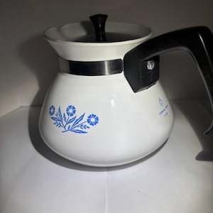 Könnte beinhalten: Weiße Vintage-Kaffeekanne mit schwarzem Griff und Deckel. Die Kanne hat ein blaues Blumenmuster und ein silbernes Band. Der Text "CORNING WARE 4 CUP" ist auf der Seite aufgedruckt.