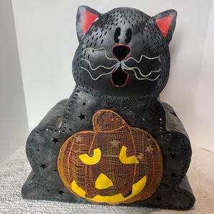 May include: Black cat-shaped Halloween decoration with a jack-o'-lantern. The cat has red ears, a stitched design, and star cutouts. The pumpkin has a mesh face and yellow accents. A festive, decorative piece.