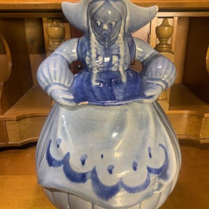 Puede incluir: Una jarra de galletas de cerámica azul y blanca con forma de niña holandesa que lleva un vestido azul y blanco y un sombrero azul. La jarra de galletas tiene un diseño floral azul y blanco en la falda.