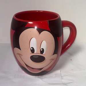May include: Red ceramic mug featuring Mickey Mouse's face. The mug has a red interior and handle, with Mickey's face prominently displayed on the front. The design includes black ears and a smiling face.