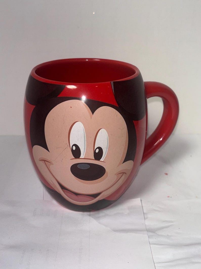 May include: Red ceramic mug featuring Mickey Mouse's face. The mug has a red interior and handle, with Mickey's face prominently displayed on the front. The design includes black ears and a smiling face.