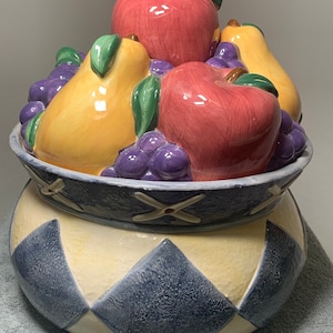 May include: Ceramic cookie jar with a blue and yellow diamond pattern. The lid features a 3D design of fruit, including red apples, yellow pears, and purple grapes.