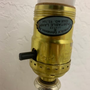 Könnte beinhalten: Nahaufnahme eines goldfarbenen Lampenfußes mit einem schwarzen Ein-/Ausschalter. Der Lampenfuß trägt ein Etikett mit der Aufschrift "Portable Lamp Listed 115V Issue No. 63 '164".