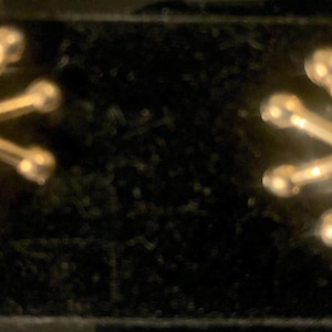 May include: A pair of gold-coloured stud earrings. Each earring has a starburst design with multiple radiating arms ending in small, round spheres. The earrings are displayed on a black surface.