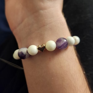 Può includere: Un braccialetto con perline bianche e viola e un ciondolo a forma di cuore dorato.