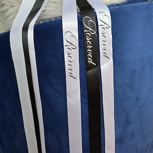 May include: Three satin ribbons, two white and one black, with the word "Reserved" in elegant script. The ribbons are draped over a blue velvet chair with silver stud detailing and a white fluffy pillow.
