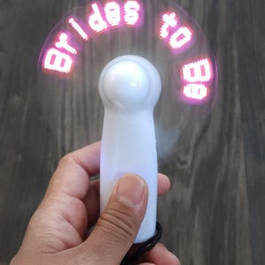 May include: A white handheld fan with the words "brides to be" displayed in pink LED lights. The fan has a rounded top and a black lanyard. The fan is held in a hand against a wood grain background.