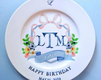May include: A white ceramic plate featuring a central blue monogram LTM, a light blue banner with the name Laken, and a pink ribbon bow. The design includes colorful floral accents, the letter L at the top, and the text HAPPY BIRTHDAY MAY 25, 2024.