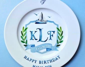 May include: A white ceramic plate featuring a blue sailboat graphic above a shield containing the monogram KLF and the name Fischer on a ribbon. The plate includes the text Happy Birthday and May 18, 2024, with green leaf accents on the sides.