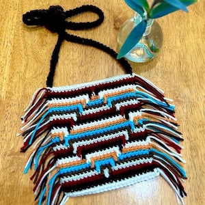 Pode incluir: Uma bolsa de crochê com um padrão geométrico em tons de branco, marrom, azul, laranja e vermelho. A bolsa tem uma alça preta e uma borda de franjas.
