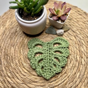 May include: A green, crocheted Monstera leaf coaster on a woven mat, with two small potted succulents behind it. The handmade coaster is a detailed item, ideal for adding a touch of nature to any home decor.