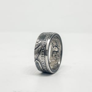 Può includere: Un anello d'argento con la scritta "UNITED STATES OF AMERICA" incisa sul lato. L'anello presenta un design dettagliato, forse da una moneta, ed è posto su uno sfondo bianco. Un gioiello unico.