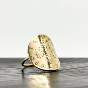 May include: A gold-colored ring with a large, oval face. The face features embossed floral designs and numbers. The ring is set against a dark, textured surface.