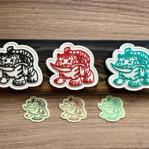 May include: A selection of tiger-shaped patches in black, red, and teal, each with a white border. Smaller, neon green and red versions are also present. The patches feature a tiger holding a blade.