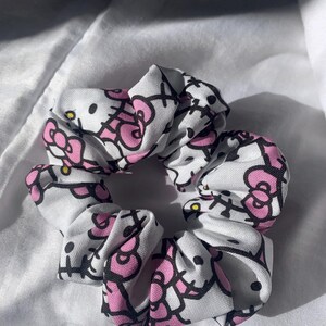 Peut inclure: Un chouchou blanc avec un motif répété d'un chat de dessin animé avec un nœud rose et des contours noirs. L'accessoire pour cheveux est conçu pour attacher les cheveux.