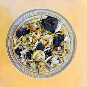 May include: A clear glass jar filled with a mix of dried herbs and berries. The contents include yellow chamomile flowers, dark purple berries, and various other dried plant parts. The jar sits on a golden-yellow surface, viewed from above.