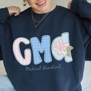 May include: Navy blue sweatshirt with the letters "GMd" in a patchwork design, featuring pink, blue, and floral accents. The words "Medical Assistant" are written below the letters in a script font. The sweatshirt is a casual style.