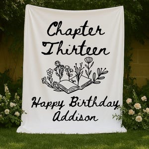 May include: A white rectangular fabric backdrop featuring black printed text that reads Chapter Thirteen and Happy Birthday Addison. Between the two lines of text is a black line art illustration of an open book with various flowers.
