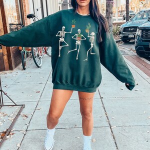May include: A dark green sweatshirt with a graphic print of three skeletons holding potted plants. The skeletons are wearing hats and have a whimsical, cartoon-like style. The sweatshirt is a crew neck style with long sleeves.