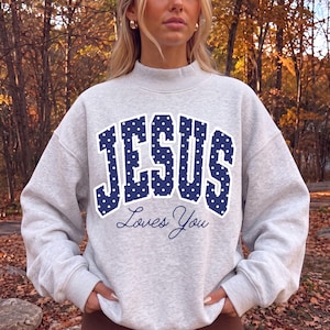 May include: A light gray crewneck sweatshirt featuring the word JESUS in large, dark blue block letters with a white polka dot pattern, positioned above the cursive text Loves You in dark blue. The garment has long sleeves and a ribbed collar.