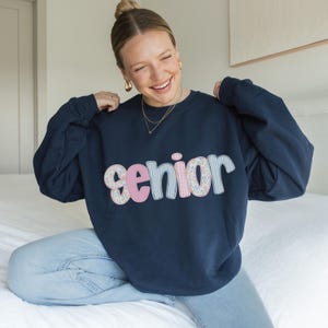 May include: Navy blue sweatshirt with the word "senior" in a patchwork design of pink, blue, and floral patterns. The sweatshirt is a crew neck style and is being held up by a person.
