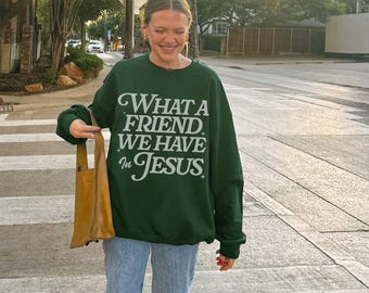 May include: A dark green sweatshirt with the text "WHAT A FRIEND WE HAVE in JESUS" in white script. The sweatshirt is paired with light blue jeans. A tan tote bag is held in the left hand.