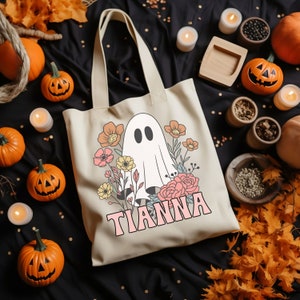 May include: A beige tote bag with a ghost and floral design. The bag has the name "TIANNA" printed on it in pink letters.