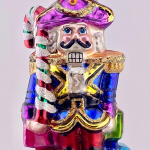 May include: A colorful glass Christmas ornament depicting a nutcracker soldier holding a candy cane. The nutcracker is wearing a purple hat, blue jacket, and red pants. The ornament is decorated with glitter and gold accents.