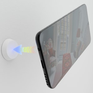 Może przedstawiać: Czarny smartfon z odblaskowym ekranem umieszczony jest w pobliżu białego, okrągłego urządzenia na białej ścianie. Wielokolorowa strzałka wychodzi z urządzenia w kierunku telefonu, sugerując połączenie bezprzewodowe lub transfer danych.